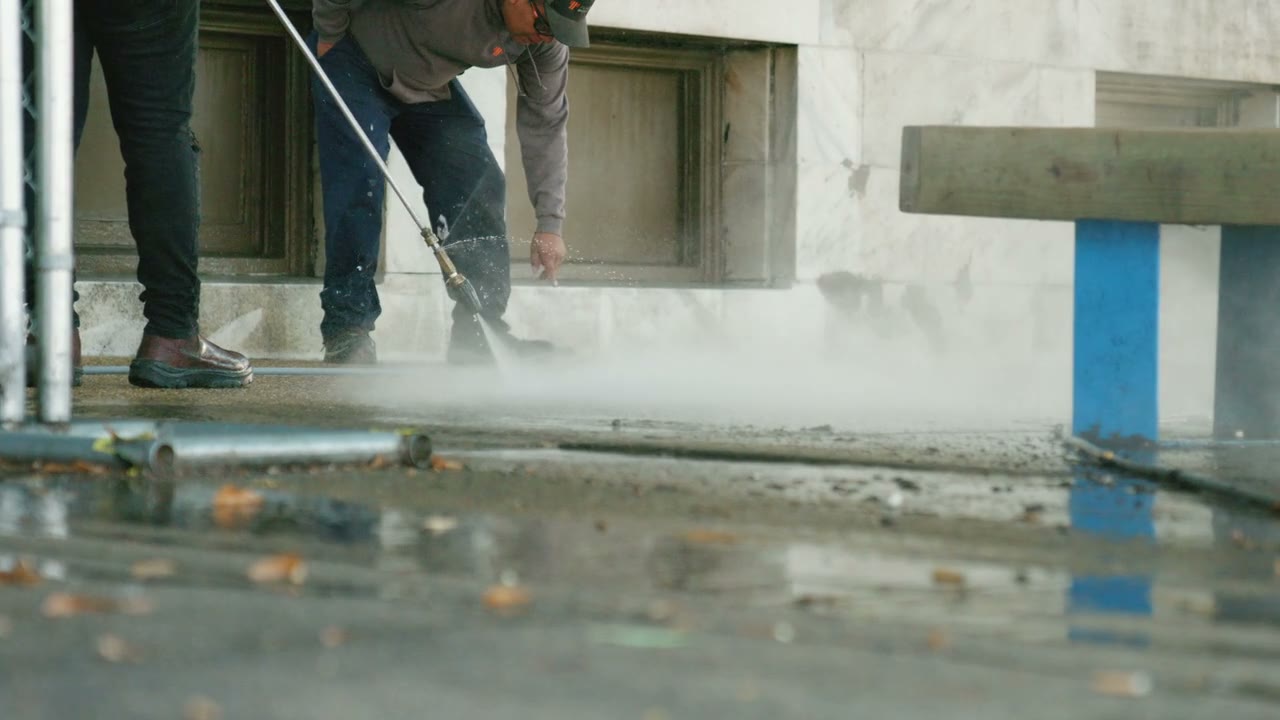 A technician pressure washing a commercial concrete surface, the spray lifting grime off the pavement